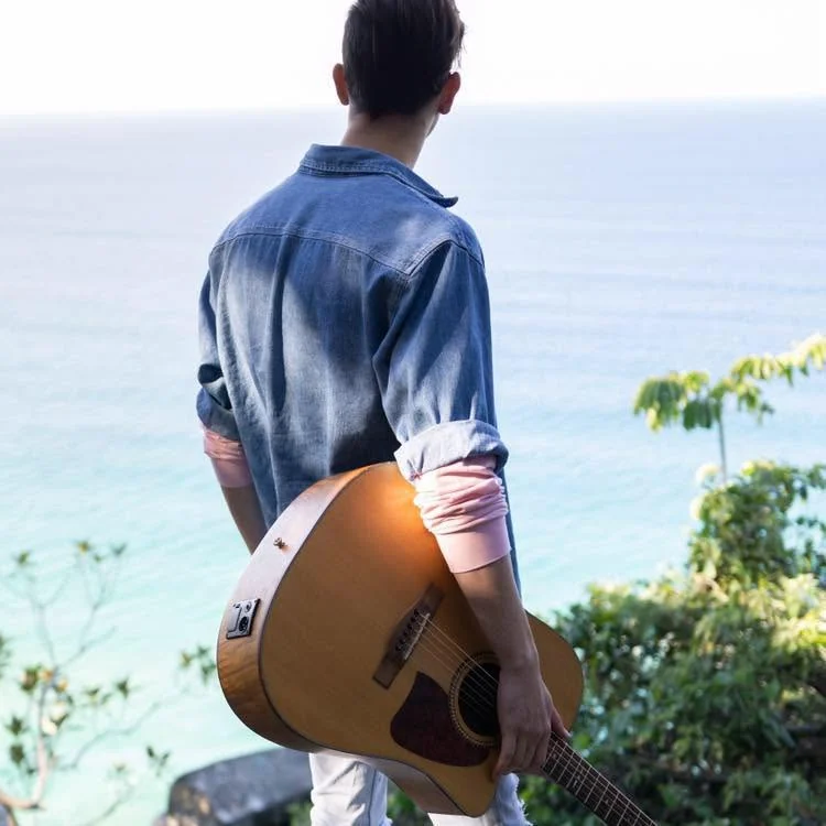Jacob Muisi holding an acoustic guitar at a coastal lookout above the ocean