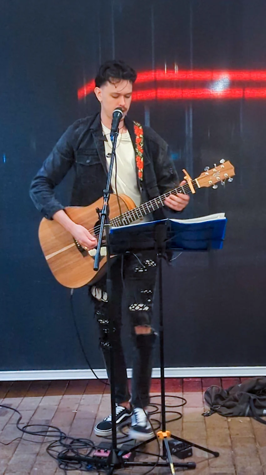 Jacob Muisi singing and playing acoustic guitar on a tiled patio, with a music stand and microphone