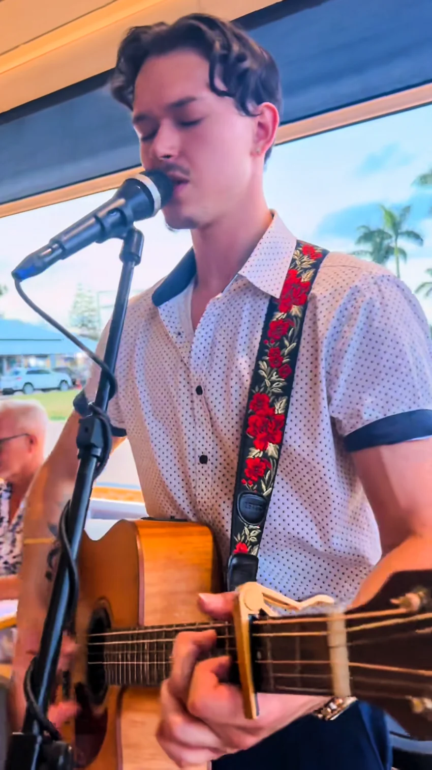 Close-up of Jacob Muisi singing into a microphone while strumming an acoustic guitar with a floral strap
