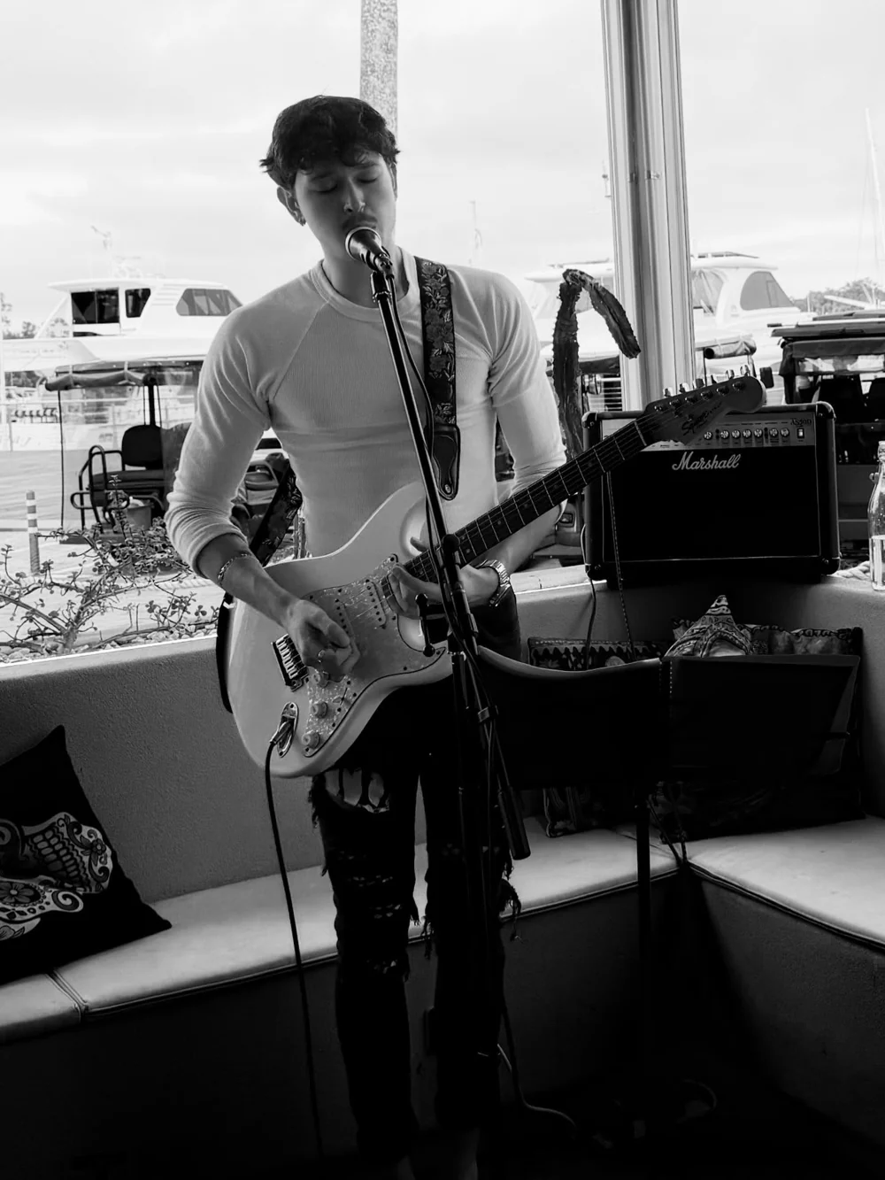 Jacob Muisi singing into a microphone and playing electric guitar beside waterfront windows